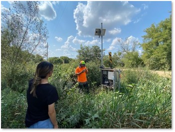 Photograph of Lala (wearing a black shirt) and another person (wearing an orange shirt) outdoors at a monitoring station by the Vermillion River.