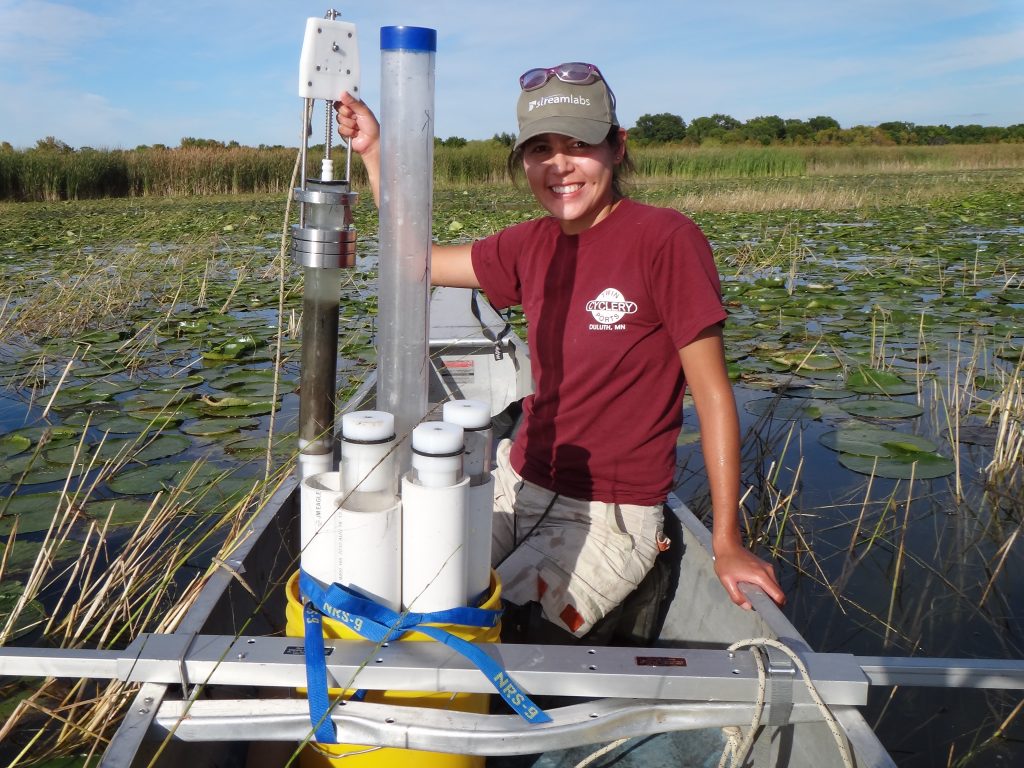 Photograph of June Sayers. She is wearing a red T-shirt and white shorts. She is in a canoe and holding a lake coring device.