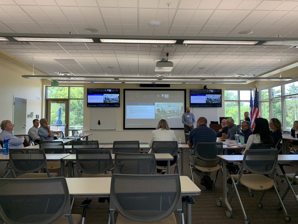 Photograph of people sitting in chairs in a meeting room. A presentation is shown on the screen in the room.