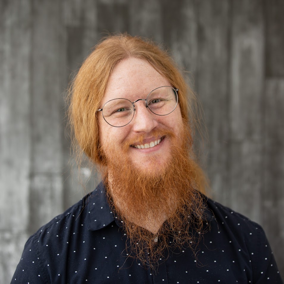 Photograph of Michael Ginsbach. Michael is wearin glasses and has red hair and a beard. He is wearing a dark blue shirt.
