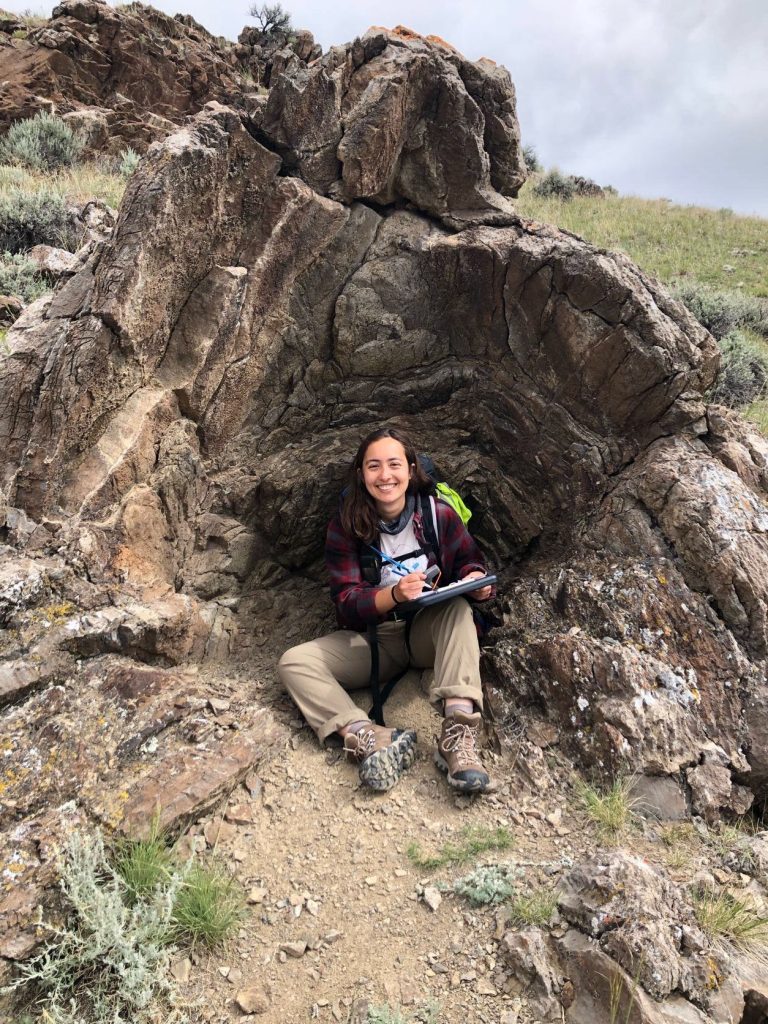 Photograph of Megan Wedal sitting by a rock formation and holding a clipboard. Megan is wearing hiking boots and khaki-colored pants.
