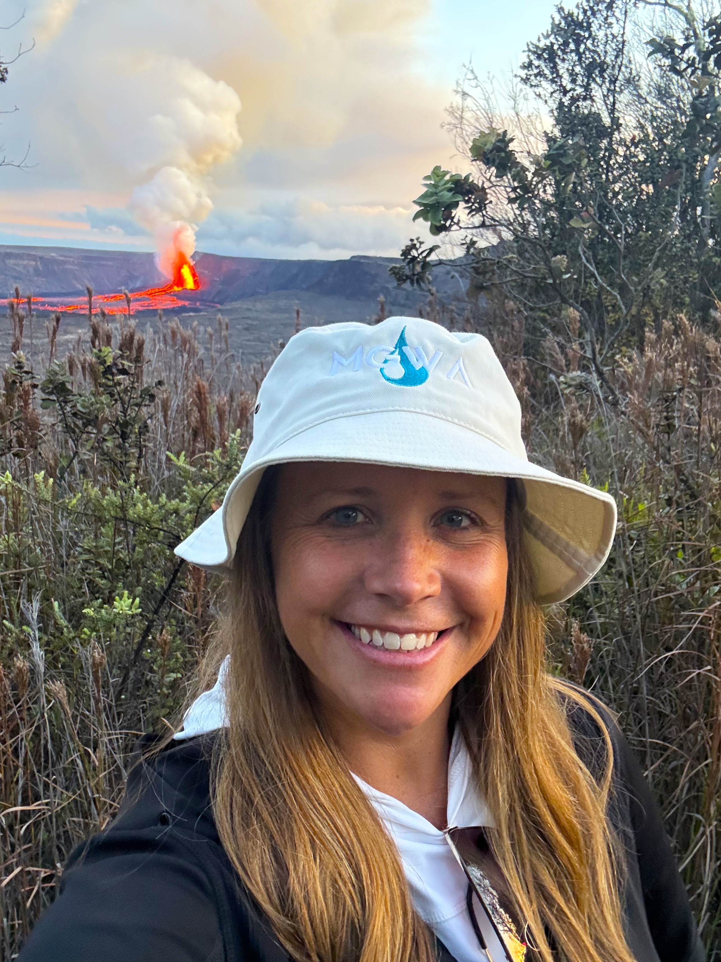 Photograph of the current MGWA President, Rebecca Higgins. Rebecca is wearing a hat with the MGWA logo, and there is an erupting volcano in the background.