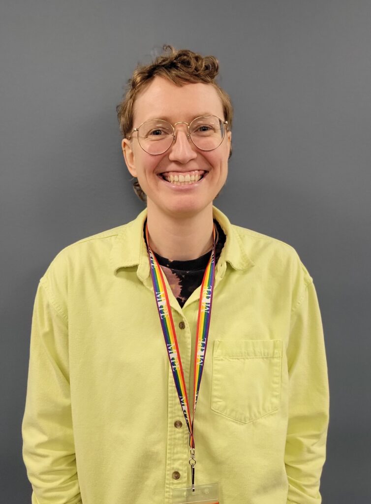 Photograph of Amanda Yourd. Amanda is smiling, wearing a yellow button-down shirt, and has short brown hair and glasses. 