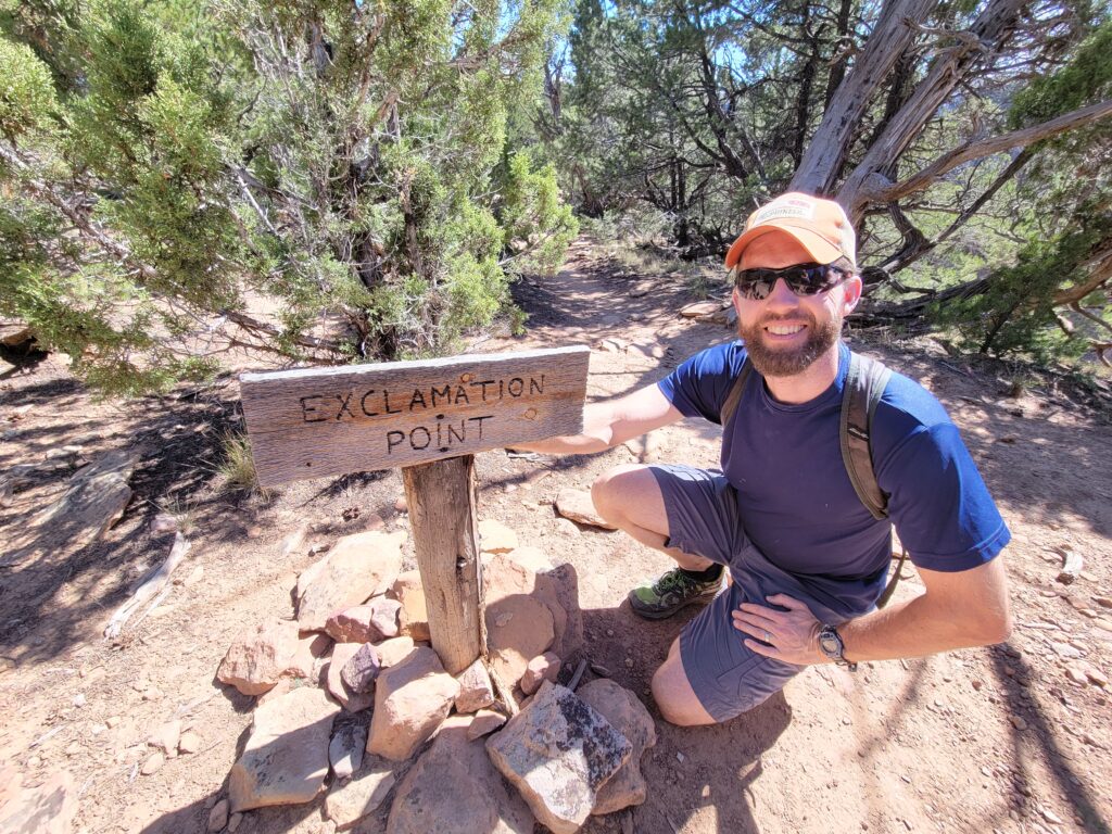 Photograph of Jared Trost. Jared is wearing an orange baseball cap, sunglasses, and a blue tee-shirt and blue shorts. He is kneeling by a wooden sign which has the words, "Exclamation point" carved into it.
