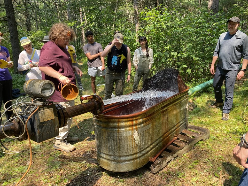 Photo of University of Minnesota Hydrogeology Field Camp attendees watching an aquifer test. A group of nine people is watching water discharge from a well into a large corrugated metal tub.