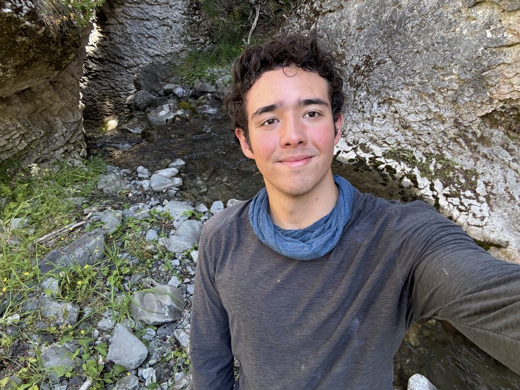 Photo of Thomas Case, recipient of 2026 Hydrogeology Field Camp Award. Thomas is wearing a gray shirt and a blue bandana and is standing in front of a bedrock outcrop.